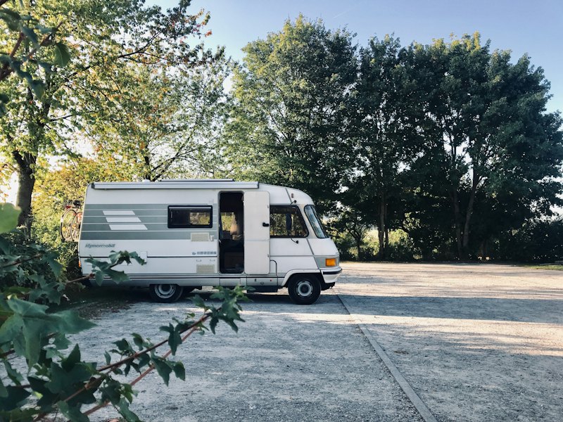 An old white motorhome parked on a gravel car park at the edge of a German forest